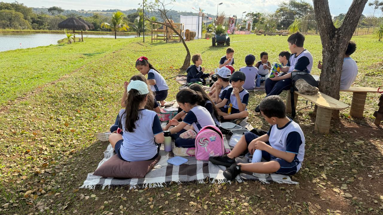 Alunos fazendo piquenique sobre uma toalha perto do lago